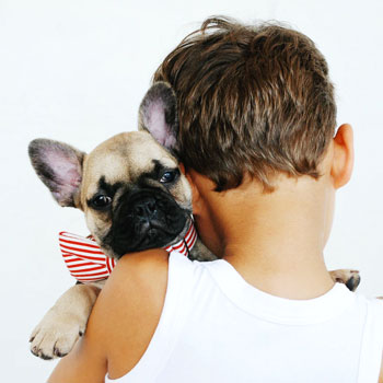 Niño de espaldas con poera blanca abrazado con su mascota un perro pequeño con una corbata roja con lineas blancas mirando a la camara